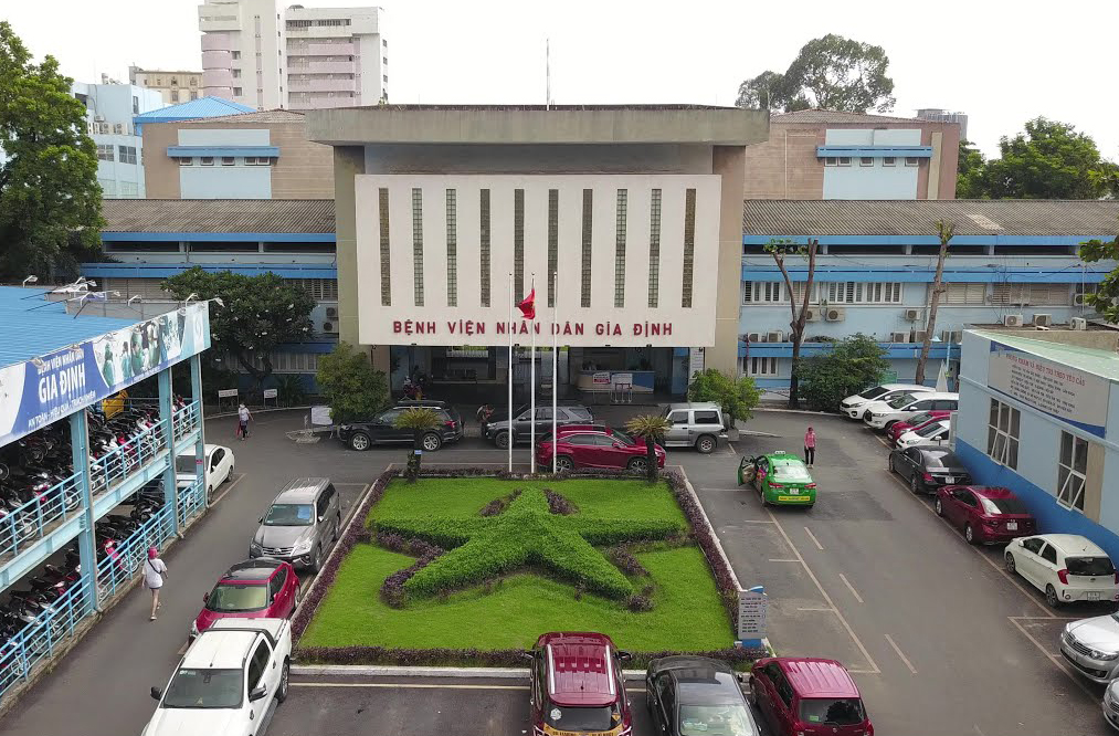 Main entrance of Nhan Dan Gia Dinh Hospital with cars parked in front, Vietnamese flags, and a star-shaped garden in Ho Chi Minh City.