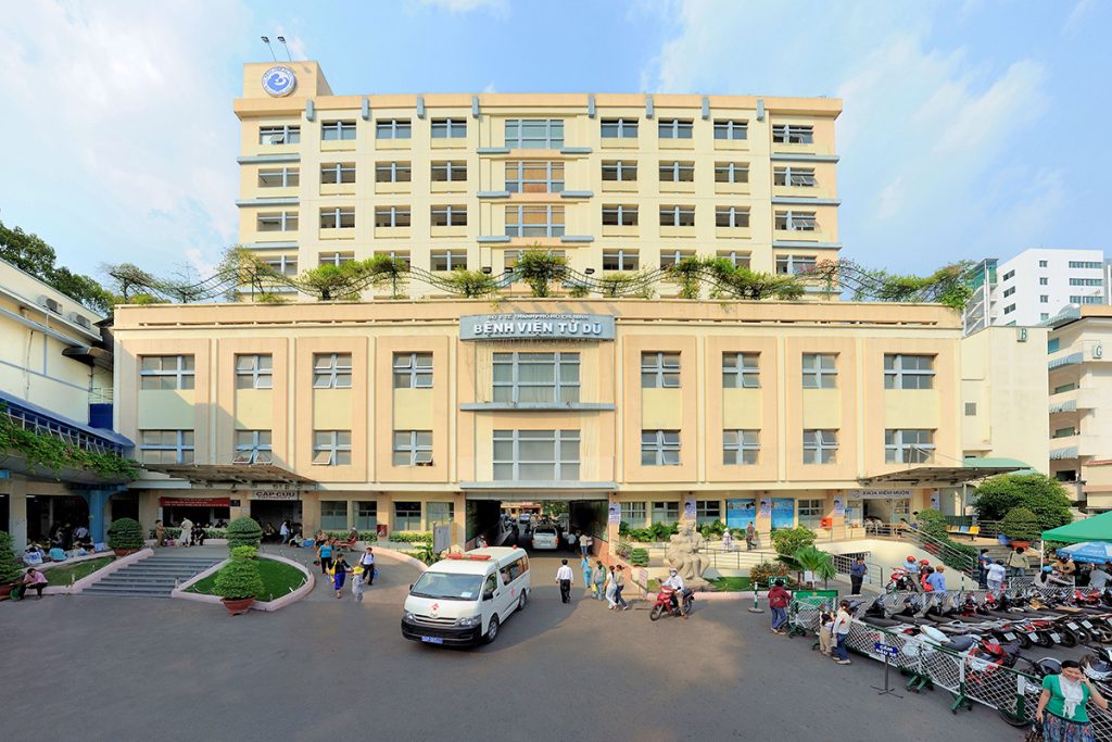 Main entrance of Tu Du Hospital in Ho Chi Minh City, showing patients, staff, and ambulances in front of the iconic yellow building.